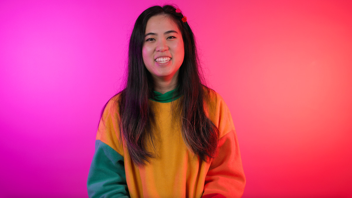 a young person with long brown hair and light brown skin grins at the camera, relaxed and happy